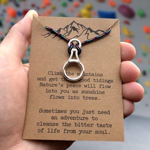 Peut inclure: Un collier pendentif argenté avec un charm en forme d'anneau, attaché à un cordon multicolore. Le pendentif est présenté sur une carte marron avec des illustrations de montagnes et du texte inspirant.