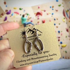 Könnte beinhalten: Silberfarbene Karabiner-Ohrringe auf einer beigen Karte mit dem Text "Especially for you - Climbing and Mountaineering Jewelry". Im Hintergrund eine Kletterwand mit farbigen Griffen und einer kletternden Person.