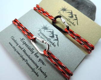 May include: Two paracord bracelets with black and red woven cord and silver and black carabiner clasps. The bracelets are displayed on a card with the text "Paracord Janka for outdoor souls" and "Especially for you - Climbing and Mountaineering Jewelry www.etsy.com/shop/Paracordjanka"