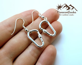 May include: Silver-colored carabiner-shaped earrings. The earrings have a hook and a small decorative screw-like element in the center. The earrings are shown against a light background. The text "ParacordJanka - for outdoor souls-" is in the upper right corner.