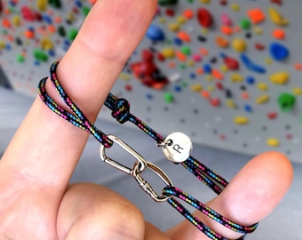 May include: A black and multi-colored rope bracelet with a silver carabiner clasp and a silver charm with the letter "R" on it.