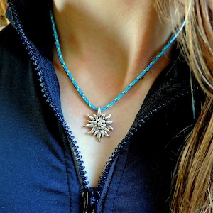 May include: A blue and green rope necklace with a silver edelweiss pendant. The necklace is worn by a person with long brown hair.