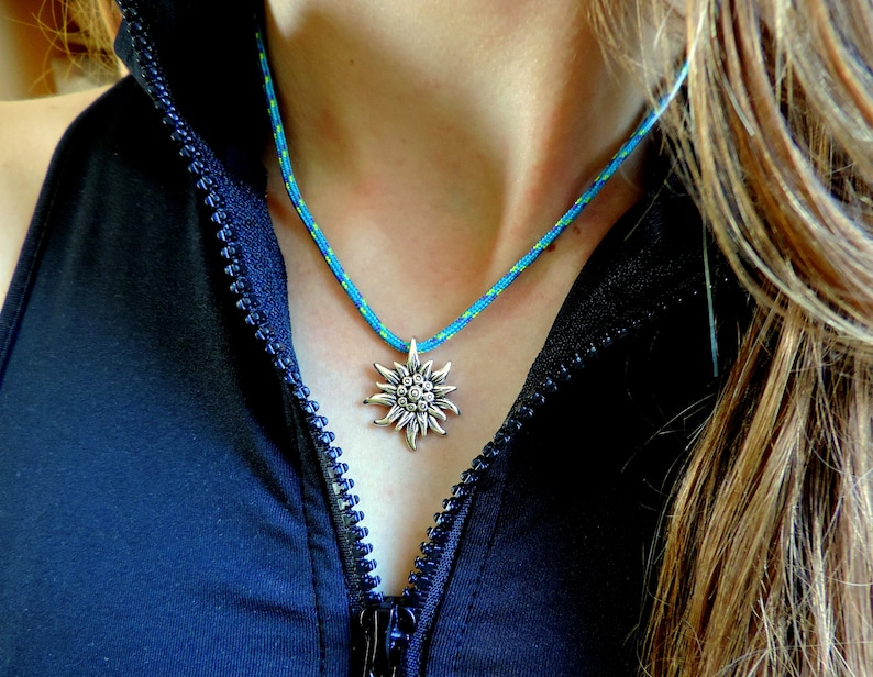 May include: A blue and green rope necklace with a silver edelweiss pendant. The necklace is worn by a person with long brown hair.