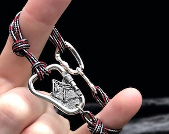 May include: Silver-toned carabiner bracelet with a black, red, and white woven cord. The carabiner features a detailed design. The bracelet is held up by a finger against a dark background. A stylish accessory.