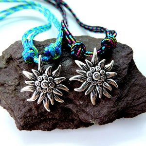 May include: Two silver Edelweiss flower pendants on blue and black braided cords. The cords are tied in knots and the pendants are hanging from the knots. The pendants are on a dark gray rock.