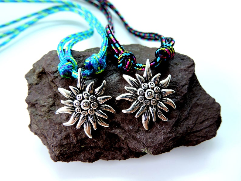 May include: Two silver Edelweiss flower pendants on blue and black braided cords. The cords are tied in knots and the pendants are hanging from the knots. The pendants are on a dark gray rock.