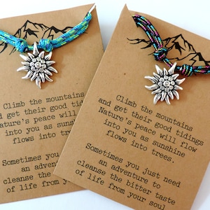 May include: Two brown cardstock tags with a mountain silhouette and a quote about climbing mountains and finding peace in nature. Each tag has a blue and green or pink and black braided cord with a silver edelweiss charm.