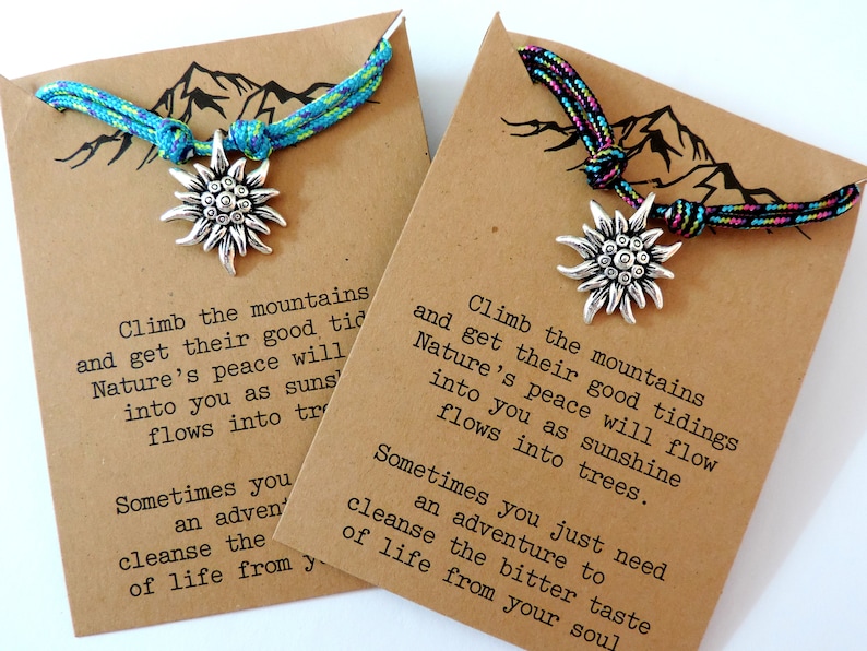 May include: Two brown cardstock tags with a mountain silhouette and a quote about climbing mountains and finding peace in nature. Each tag has a blue and green or pink and black braided cord with a silver edelweiss charm.