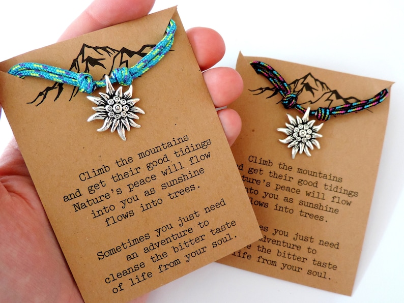 May include: Two brown cardstock tags with a mountain silhouette and a quote about climbing mountains and finding peace. Each tag has a silver charm of a stylized mountain flower attached to a colorful braided cord.