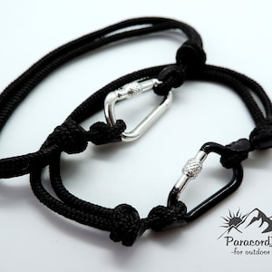 May include: Two black paracord bracelets with silver and black carabiner clasps. The bracelets are tied with a knot at each end. The text "ParacordJanka -for outdoor souls- " is visible in the lower right corner of the image.