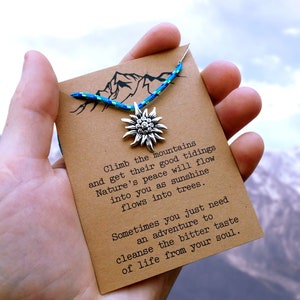 May include: A brown card with a blue and green cord and a silver charm in the shape of a flower. The card has the text "Climb the mountains and get their good tidings Nature's peace will flow into you as sunshine flows into trees. Sometimes you just need an adventure to cleanse the bitter taste of life from your soul."