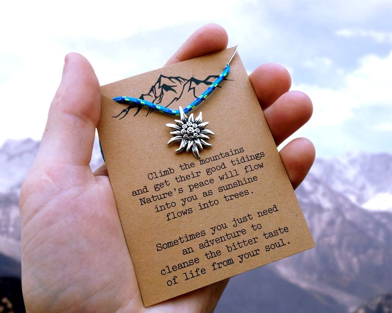 May include: A brown card with a blue and green cord and a silver charm in the shape of a flower. The card has the text "Climb the mountains and get their good tidings Nature's peace will flow into you as sunshine flows into trees. Sometimes you just need an adventure to cleanse the bitter taste of life from your soul."