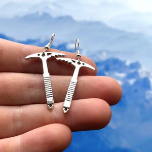 May include: A pair of silver-colored earrings shaped like miniature ice axes. Each earring features a pick, a head, and a ribbed shaft. The earrings are held in a hand against a blurred mountain backdrop.
