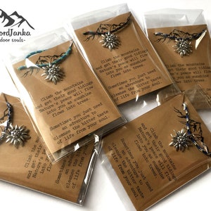 May include: Five small brown paper bags with clear plastic windows, each containing a silver charm with a mountain flower design and a blue and white paracord bracelet. The bags have a printed message that reads "Climb the mountains and get their good tidings. Nature's peace will flow into you as sunshine flows into trees. Sometimes you just need an adventure to cleanse the bitter taste of life from your soul."