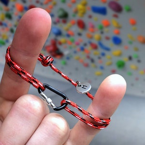 Pu&ograve; includere: Un braccialetto in corda rosso e nero con un moschettone argentato e un ciondolo argentato con la lettera "M". Il braccialetto &egrave; indossato su un dito.