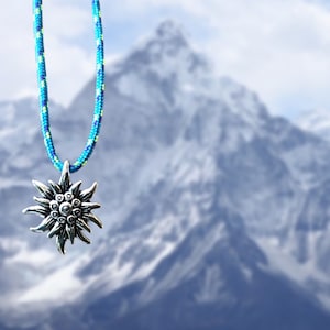 May include: A silver edelweiss pendant on a blue and green rope necklace. The pendant is hanging in front of a blurry background of a snow-capped mountain.