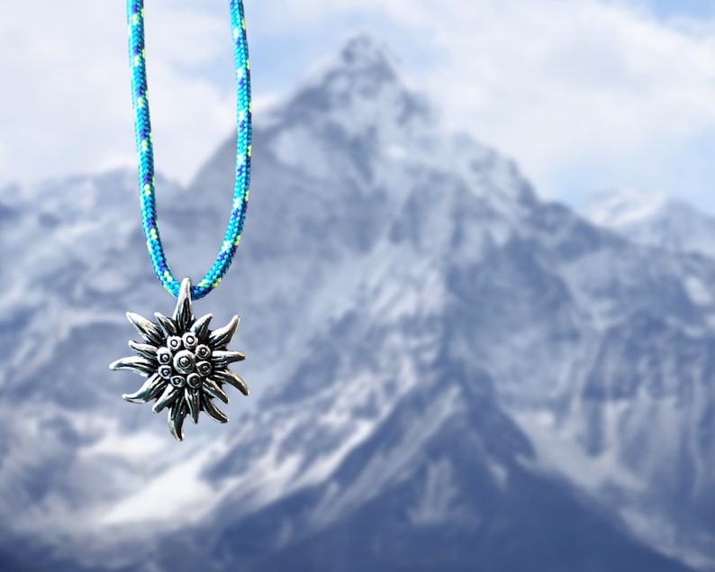 May include: A silver edelweiss pendant on a blue and green rope necklace. The pendant is hanging in front of a blurry background of a snow-capped mountain.