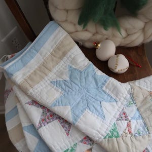 Könnte beinhalten: Ein gesteppter Weihnachtsstrumpf mit einer hellblauen Sternapplikation auf weißem Grund, ergänzt durch beige Streifen und Stoffe mit Blumenmuster. Der Strumpf ist mit sichtbaren Nähten und einem Umschlag versehen und zeigt ein Patchwork-Design.
