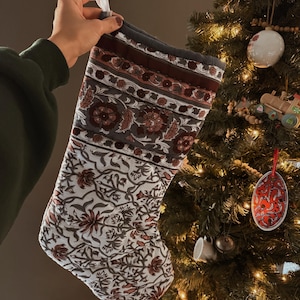 Könnte beinhalten: Ein gesteckter Weihnachtsstrumpf mit floralem Muster in Weiß, Grau und Burgunderrot. Der Strumpf hat eine Aufhängeschlaufe. Im Hintergrund ist ein geschmückter Weihnachtsbaum zu sehen.