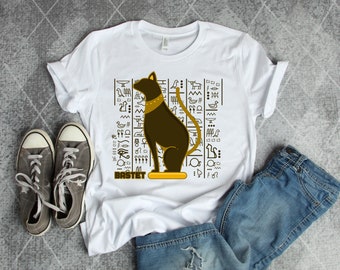 May include: White t-shirt with a black and gold illustration of the Egyptian goddess Bastet, a cat deity. The illustration is surrounded by hieroglyphs and the word "BASTET" is written below the image.