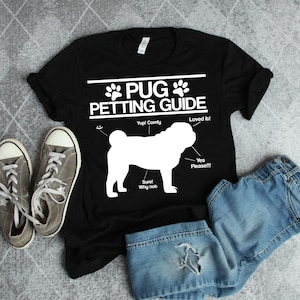 May include: Black t-shirt with a white pug silhouette and the text "PUG PETTING GUIDE" with instructions on how to pet a pug. The shirt is laid out on a gray surface with a pair of gray sneakers and blue jeans.