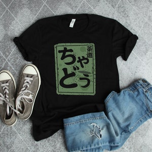 May include: Black t-shirt with a green rectangular graphic featuring Japanese characters. The shirt is paired with gray sneakers and denim shorts. The graphic reads "茶道" and "ちやとう".