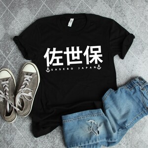 May include: Black t-shirt with white text that says "佐世保" and "Sasebo Japan" with an anchor on either side of the text. The shirt is laying on a gray surface with a pair of gray sneakers and blue jeans.