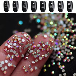 May include: A close-up of a variety of iridescent rhinestones in different sizes. The rhinestones are scattered on a finger and a black surface. The sizes of the rhinestones are labelled with the text "SS3 1.3MM", "SS4 1.5MM", "SS5 1.7MM", "SS6 1.9MM", "SS8 2.4MM", "SS10 2.7MM", "SS12 3.0MM", and "SS16 3.8MM".
