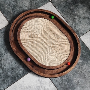 Könnte beinhalten: Ein ovales Tablett aus Holz mit einem Einsatz aus geflochtenem Seil. Das Tablett hat eine natürliche Holzoptik und ist mit drei bunten Kugeln verziert.