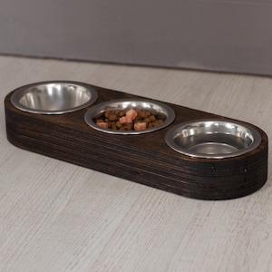 May include: A dark brown wooden pet food bowl stand with three stainless steel bowls. The stand is on a light wood surface. One bowl has a small amount of brown and pink pet food.