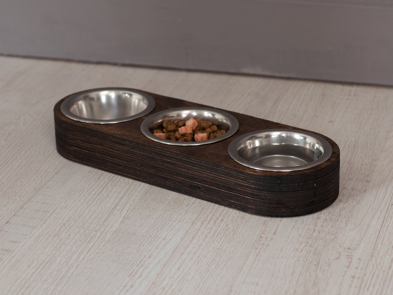 May include: A dark brown wooden pet food bowl stand with three stainless steel bowls. The stand is on a light wood surface. One bowl has a small amount of brown and pink pet food.