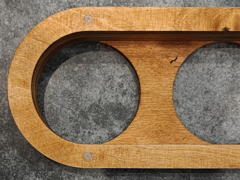 May include: A wooden coaster with two circular cutouts for holding mugs or glasses. The coaster has a natural wood finish and is decorated with two small silver dots.