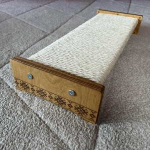 Op de afbeelding: Een krabplank voor katten gemaakt van natuurlijk hout en gevlochten touw. De houten uiteinden zijn versierd met een geëtst geometrisch patroon en vastgezet met metalen schroeven. De plank is ontworpen voor katten om aan te krabben en hun nagels te slijpen.