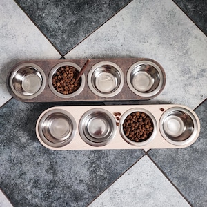 May include: Two wooden pet food bowls with four stainless steel bowls. The top bowl is dark brown wood and the bottom bowl is light wood. Both bowls have dry pet food in one of the bowls.