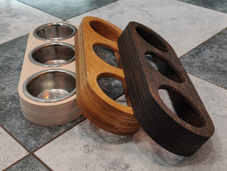 May include: Three wooden bowls with different finishes. The bowl on the left is white with three round indentations. The middle bowl is light brown with two round indentations. The bowl on the right is dark brown with three round indentations.