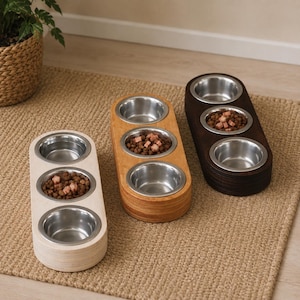 Peut inclure: Trois stations d'alimentation pour animaux de forme allongée, chacune dotée de trois bols circulaires en acier inoxydable insérés dans une base en bois. Les bases présentent des tons de bois clair, moyen et foncé, avec des croquettes.