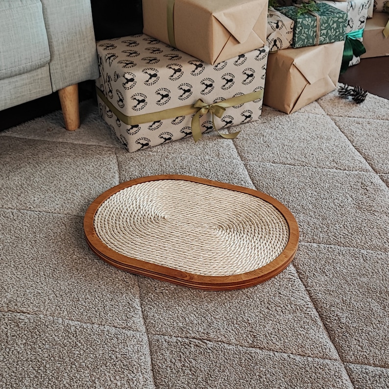 May include: A round, oval-shaped cat scratching post with a natural rope surface and a wooden frame. The scratching post is on a gray carpet.