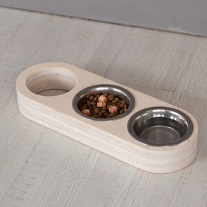 May include: A wooden pet food bowl stand with three stainless steel bowls. One bowl is filled with brown pet food and orange treats.