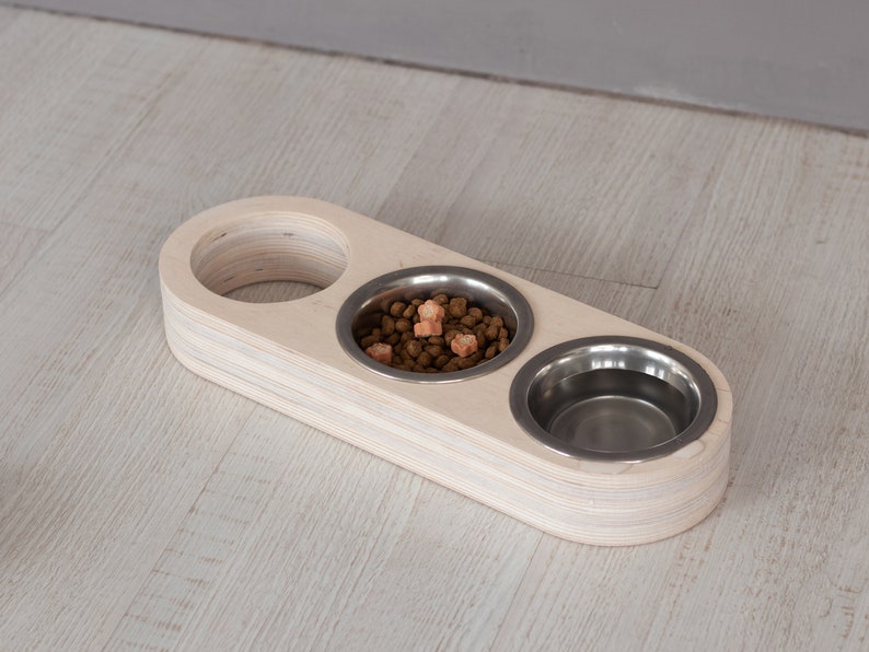 May include: A wooden pet food bowl stand with three stainless steel bowls. One bowl is filled with brown pet food and orange treats.