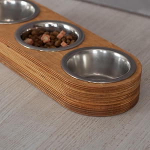 May include: A wooden pet food bowl stand with three stainless steel bowls. The stand is made of light brown wood and has a rounded shape. The bowls are empty.