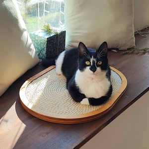 May include: A black and white cat sits on a round, natural fiber scratching post with a wooden frame. The scratching post is on a wooden surface.