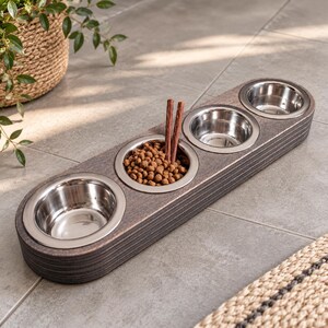 May include: A dark brown, elongated wooden pet feeder featuring three circular cutouts, each holding a removable stainless steel bowl. The center bowl is filled with brown dry pet food and two brown stick-shaped treats.