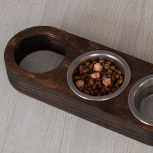 May include: A dark brown wooden pet food bowl holder with three stainless steel bowls. One bowl is filled with brown kibble and pink flower-shaped treats.
