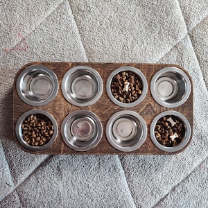 May include: A wooden dog bowl holder with eight stainless steel bowls. Some bowls are filled with brown kibble and bone-shaped treats. The holder is a dark brown colour and sits on a grey surface.