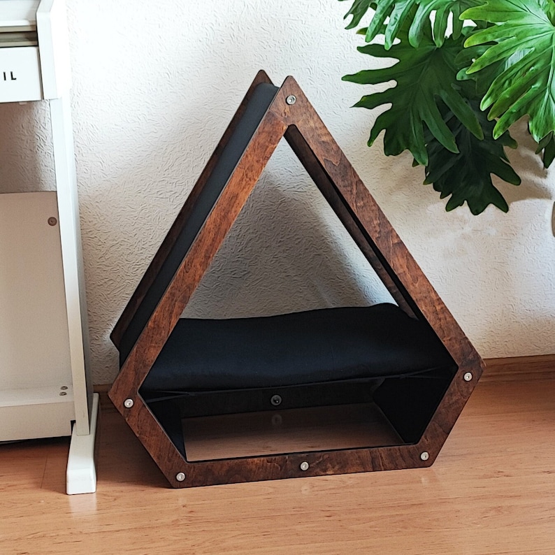 May include: A wooden pet bed shaped like a triangle with a black cushion inside. The bed is made of dark wood with a natural finish and has metal accents.