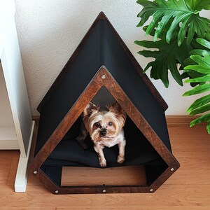 May include: A brown wooden dog house with a black fabric interior. The dog house has a triangular shape with a pointed roof. A small brown and white dog is sitting inside the house.