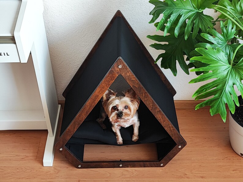 May include: A brown wooden dog house with a black fabric interior. The dog house has a triangular shape with a pointed roof. A small brown and white dog is sitting inside the house.