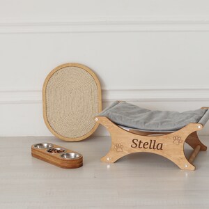 May include: A wooden pet bed with a gray cushion and a paw print design. The bed is engraved with the name "Stella". A wooden scratching post and a triple bowl feeder are also shown.
