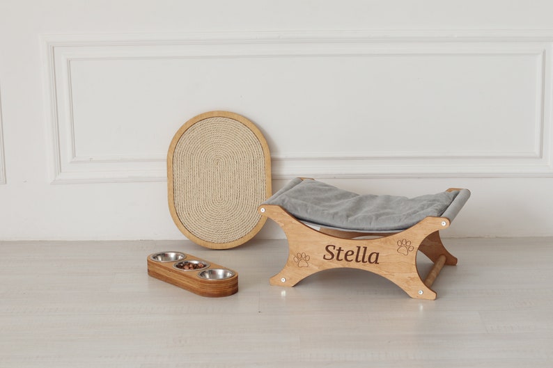 May include: A wooden pet bed with a gray cushion and a paw print design. The bed is engraved with the name "Stella". A wooden scratching post and a triple bowl feeder are also shown.