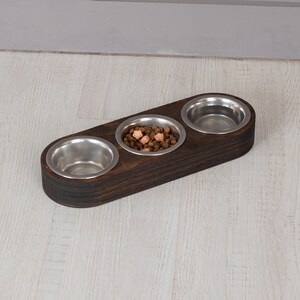 May include: A dark brown wooden pet food bowl with three stainless steel bowls. The center bowl has dry pet food in it.